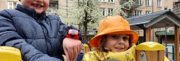 Två förskolebarn varav en med en röd rörelsemätare på handleden, ler mot kameran på en lekplats utomhus