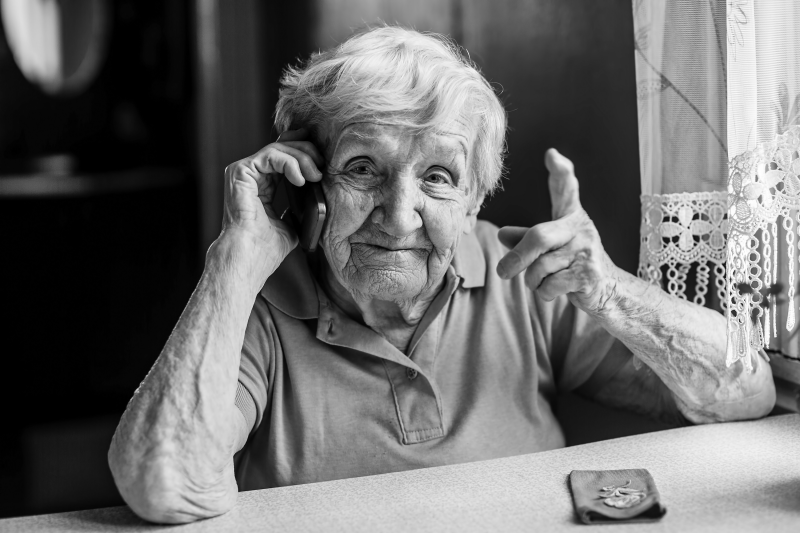 Äldre kvinna sitter vid ett bord och talar i en mobiltelefon. Foto: AdobeStock