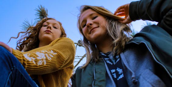Två flickor tittar in i kameran och ler, i bakgrunden syns en blå himmel.