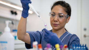 En person i vårdkläder och skyddsglasögon drar med en pipett upp vätska ur ett provrår.