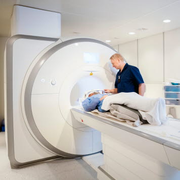 En patient ligger på en brits till en magnetröntgenmaskin. En vårdgivare står bredvid. Foto: AdobeStock