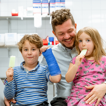 En vuxen och två barn sitter på en brits. Det ena barnet har en gipsad arm. Båda barnen äter glass. Foto: Yanan Li