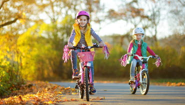 Två flickor cyklar på en väg kantad av träd och buskar med bebyggelse i bakgrunden,