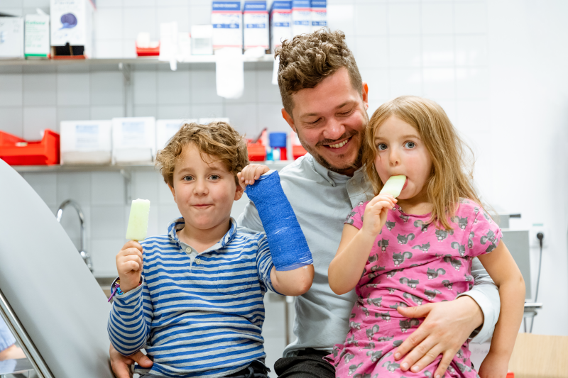 En vuxen och två barn sitter på en brits. Det ena barnet har en gipsad arm. Båda barnen äter glass. Foto: Yanan Li