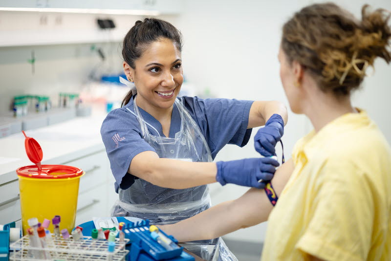 Ett laboratorium där en person i vårdkläder sätter fast ett spännband inför blodprovtagning på en person i privata kläder.