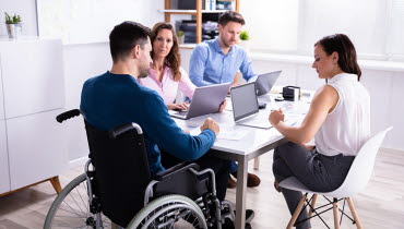 MAn med funktionsnedättning sitter i en rullstol vid ett mötesbord tillsammans med kollegor.
