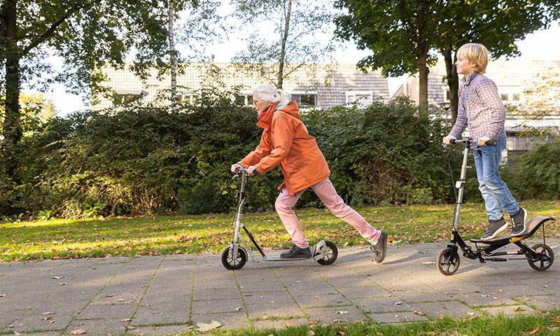 Pojke och äldre kvinna cyklar sparkcykel på en trottoar i ett grönskande bostadsområde