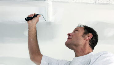Målare som målar ett tak i en obekväm arbetsställning med högt lyft arm.